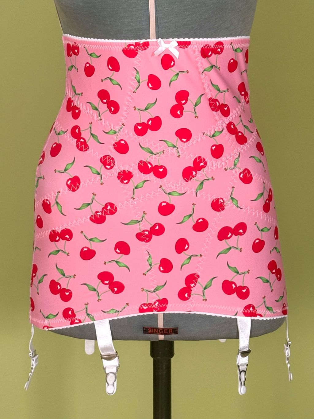 Pink girdle with a red cherry print, displayed on a gray dress form mannequin against a solid olive green background.
