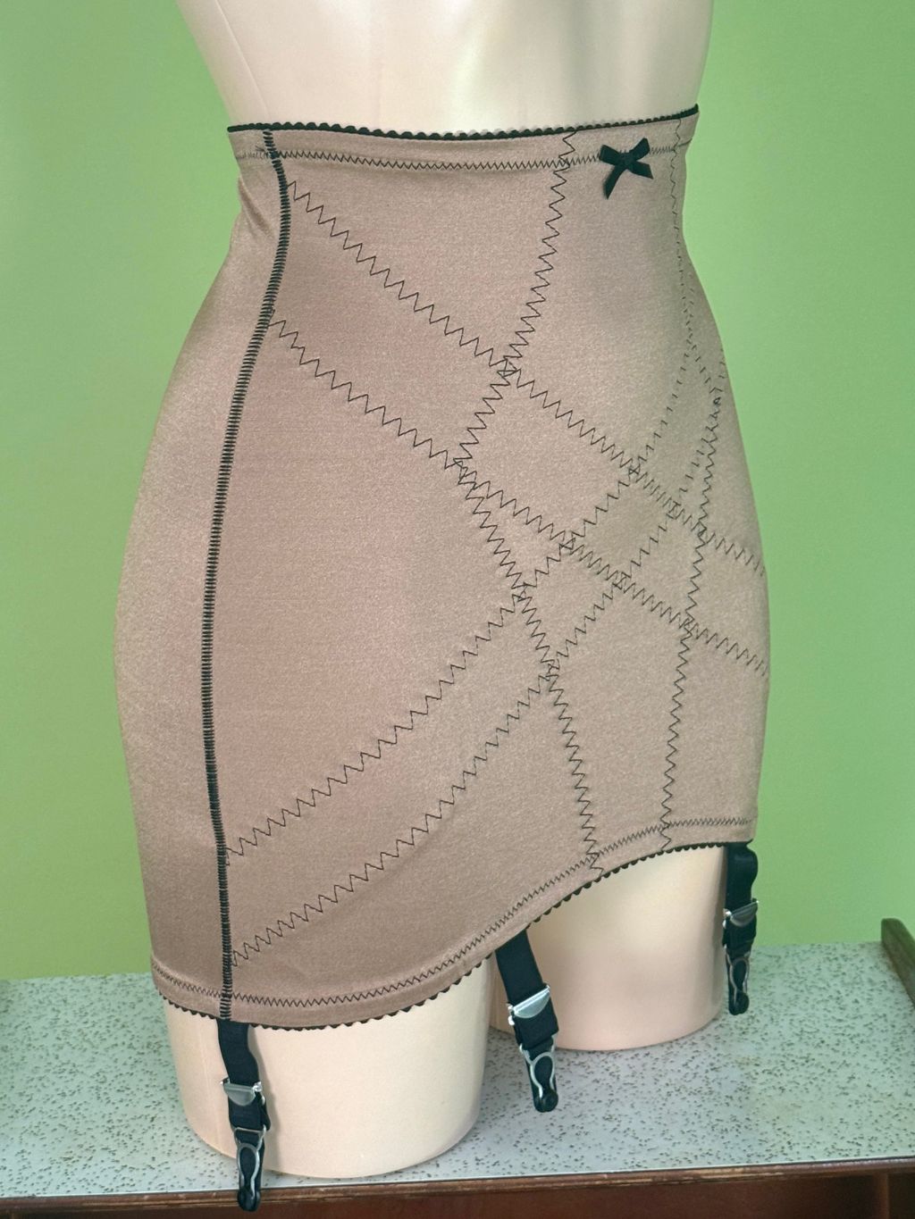 A beige high-waisted Girdle with zigzag stitching and a small black bow at the center front, displayed on a mannequin with attached black garter straps against a light green background.