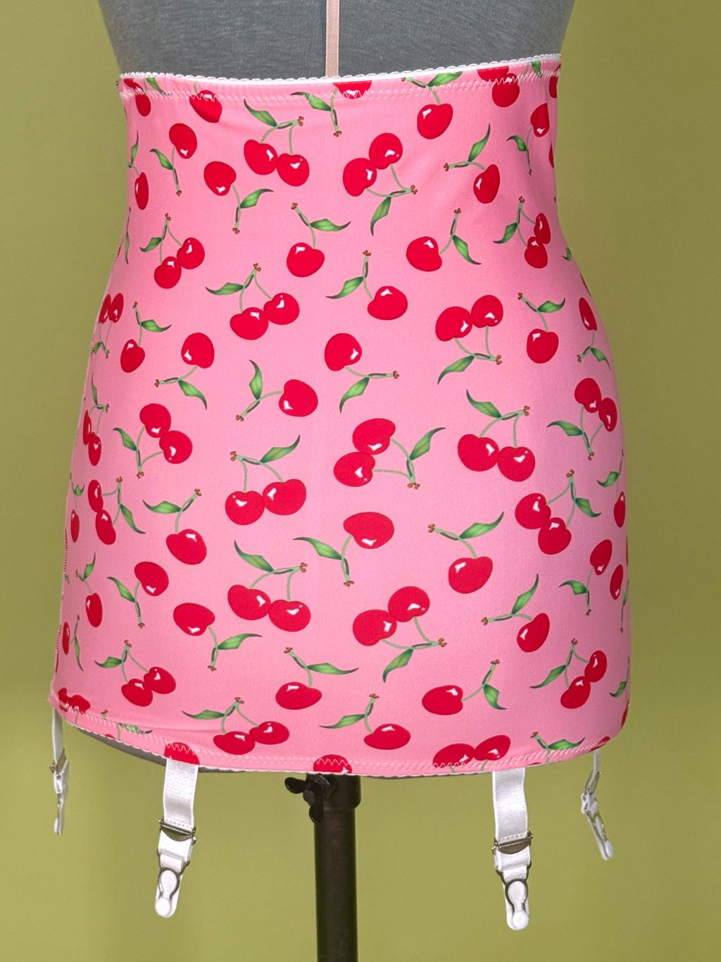 Pink girdle with a red cherry print, displayed on a mannequin with white garter straps hanging from the bottom, against a solid olive green background.