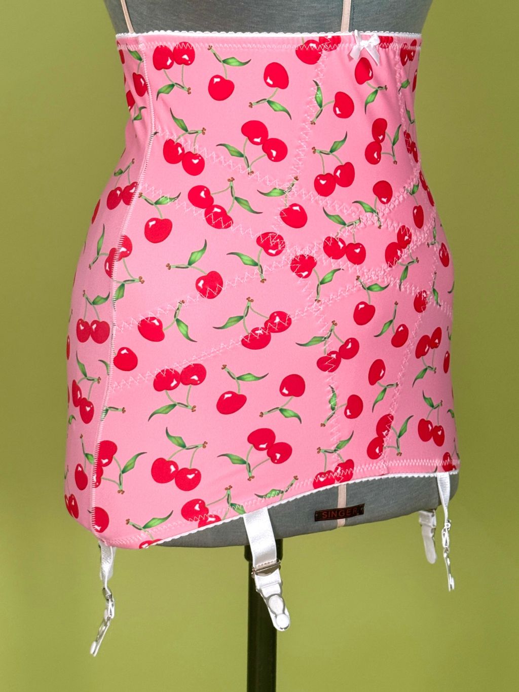 Pink girdle with a red cherry print, displayed on a gray dress form against an olive green background.