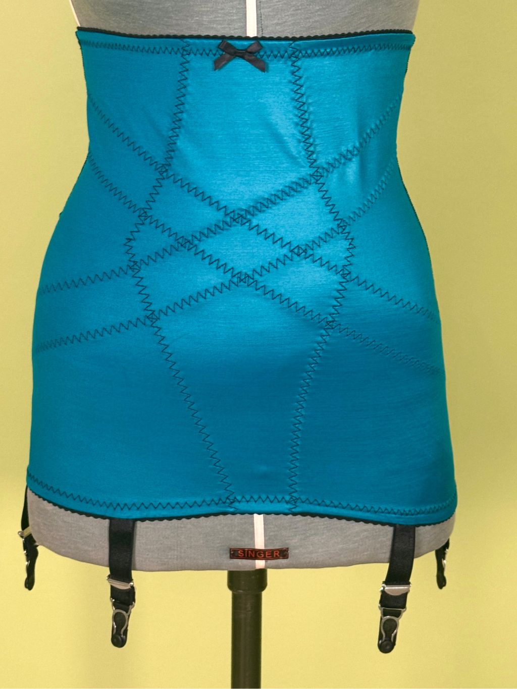 A bright turquoise blue girdle with diagonal contrast stitching, a small black bow at the top, and black garter straps hanging from the bottom, displayed on a gray dress form against a pale yellow background.