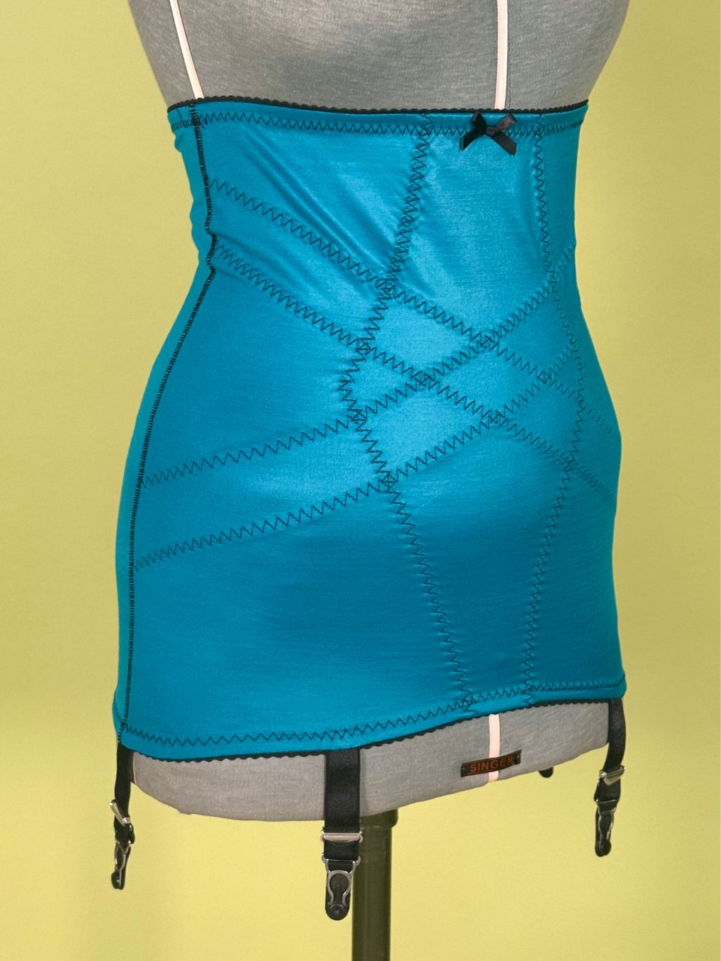 A bright turquoise blue girdle with diagonal contrast stitching, black garter straps, and a small black bow at the top, displayed on a gray dress form against a pale yellow background.