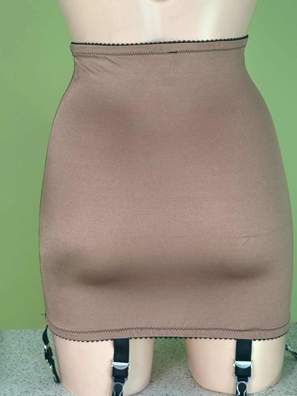 A beige high-waisted shapewear Girdle displayed on a mannequin, featuring black garter straps hanging from the hem against a light green background.
