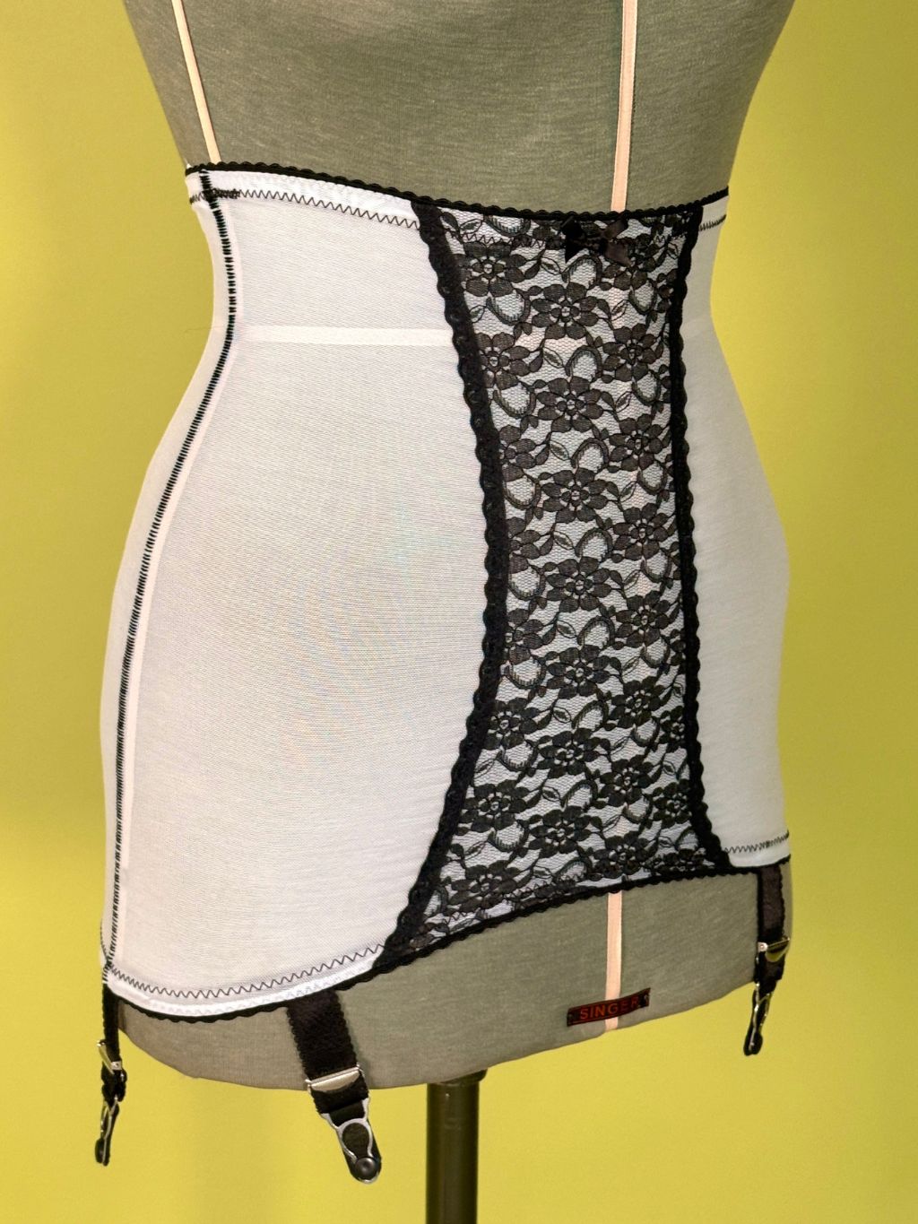 Cream girdle with black lace panel and black garter straps, displayed on a gray dress form against a yellow background.