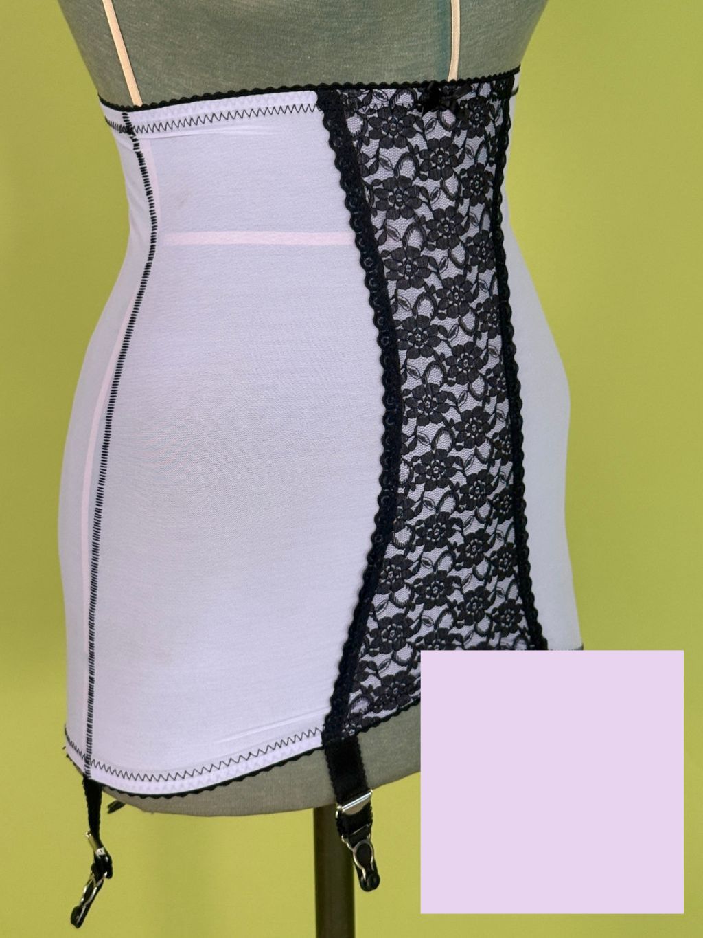 Lilac girdle with black lace panel and black garter straps, displayed on a torso mannequin against a yellow-green background. A lavender color swatch box is overlaid in the bottom right corner.