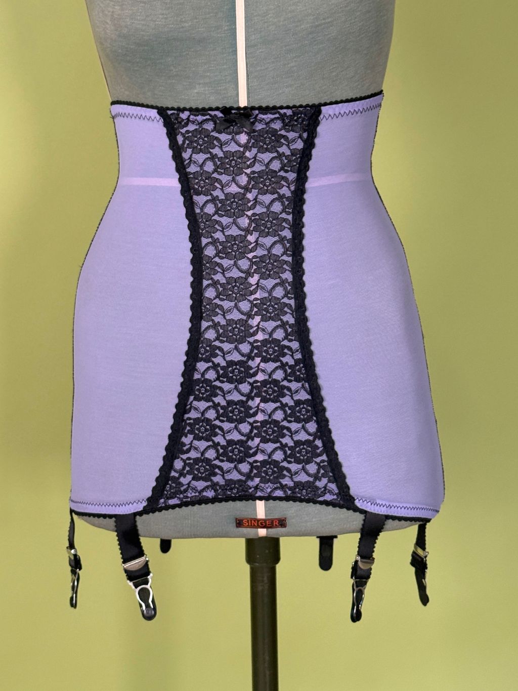 A lavender and black girdle displayed on a gray dress form against a pale yellow-green background.
