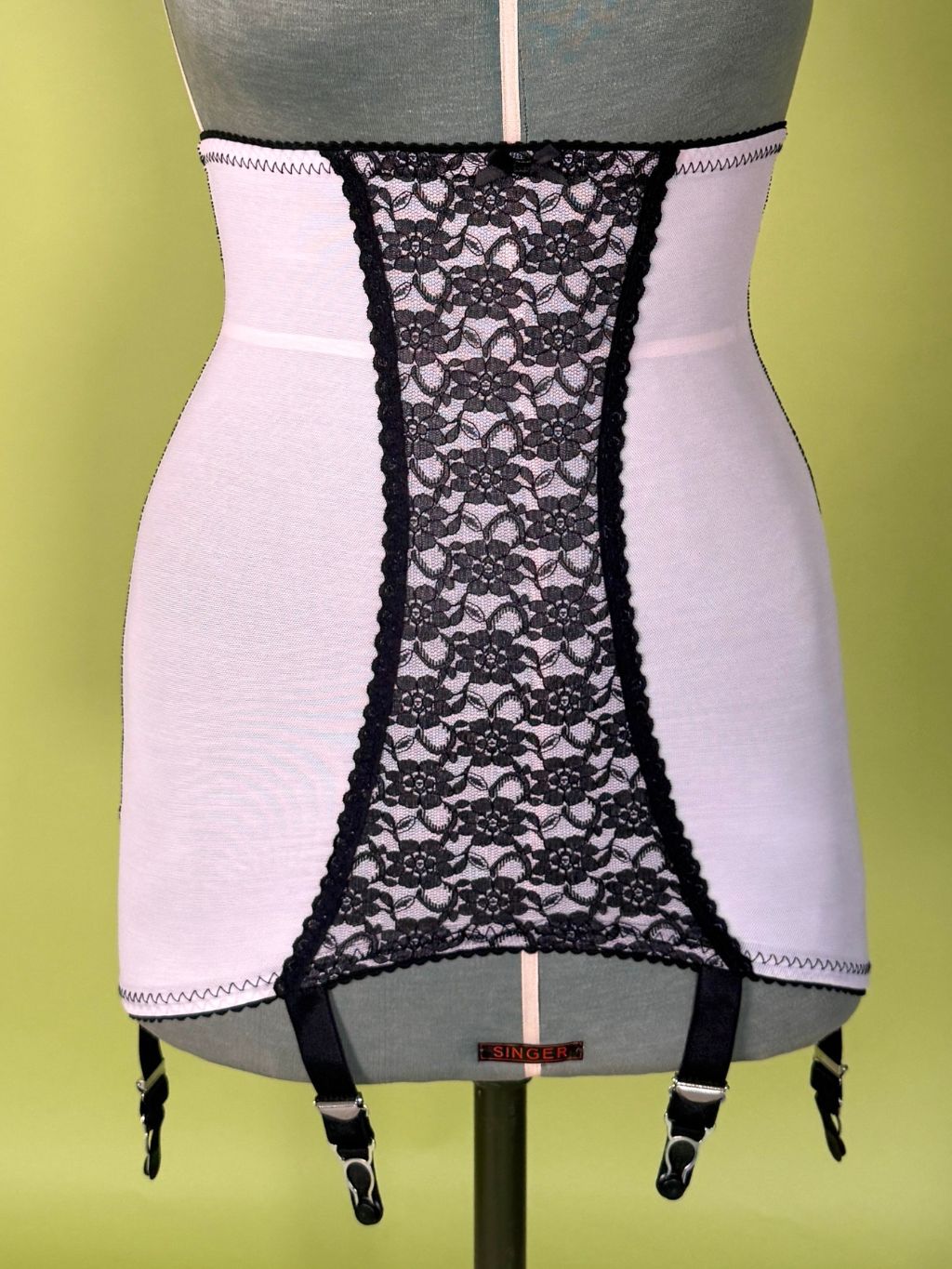 Pink girdle with black lace center panel and black garter straps, displayed on a gray dress form against a yellow-green background.