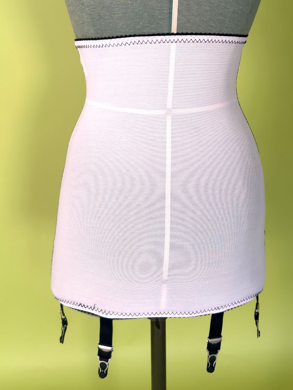 Pink girdle with black garter straps, displayed on a gray dress form mannequin against a yellow-green background.