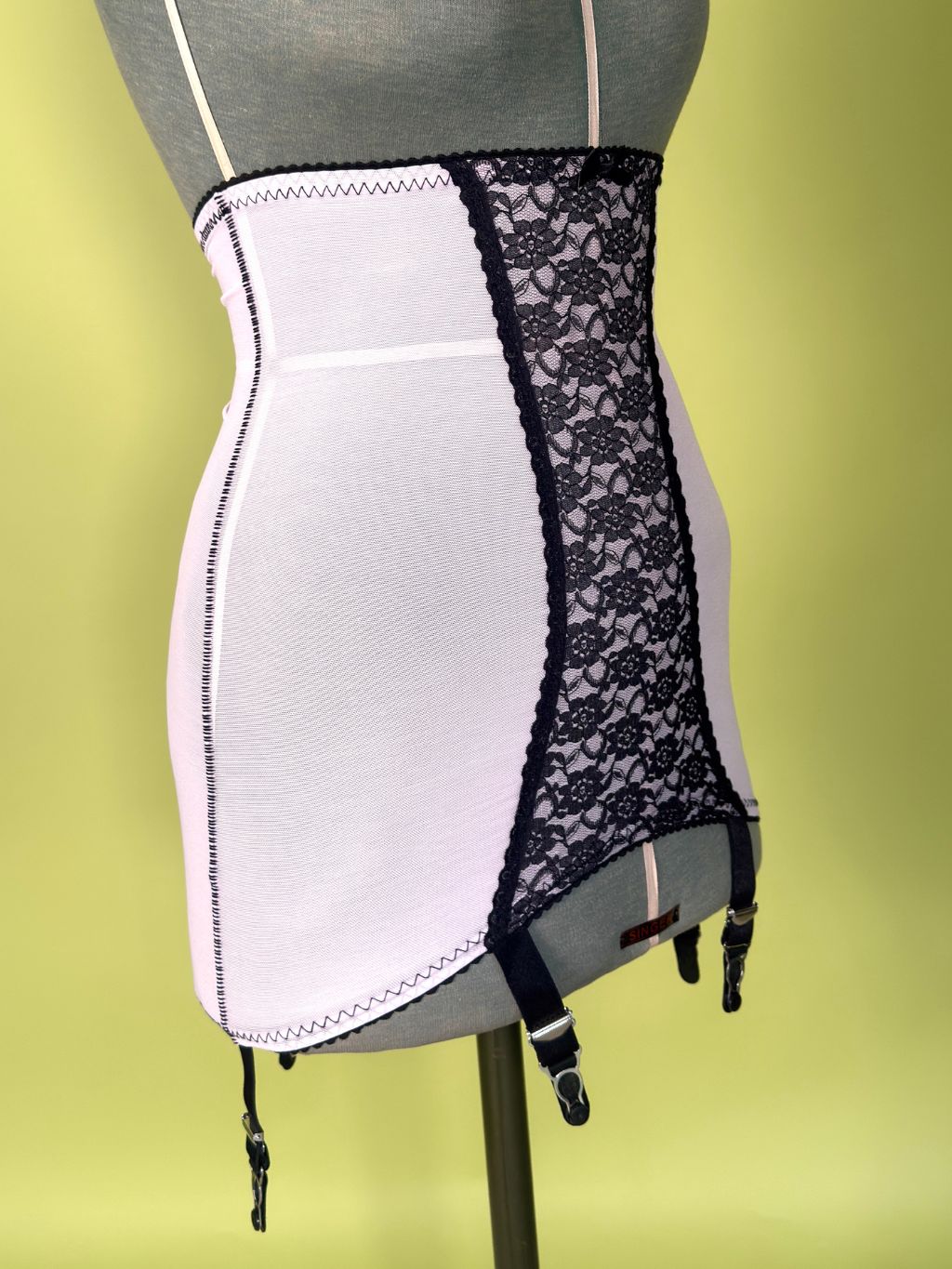 A pink and black girdle with black lace panels and garter straps, displayed on a gray mannequin torso against a yellow-green background.