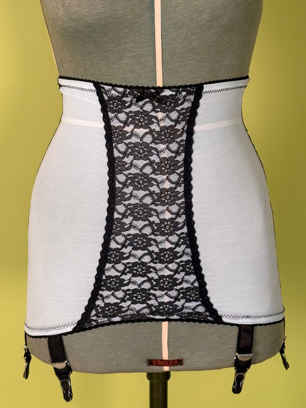Cream girdle with black lace center panel and black garter clips, displayed on a gray dress form against a yellow background.