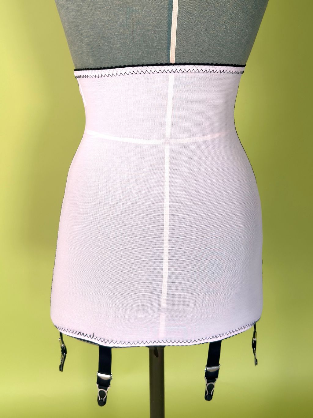 Cream girdle  displayed on a dress form mannequin, with black garter straps hanging from the bottom against a yellow-green gradient background.