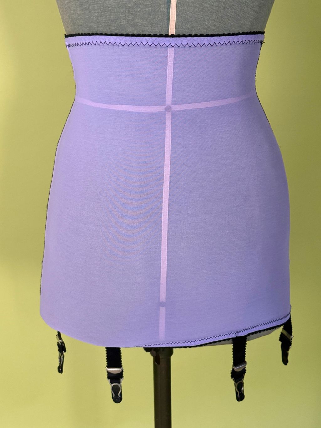 A lavender purple girdle with garter straps, displayed on a gray dress form mannequin against a solid yellow-green background.