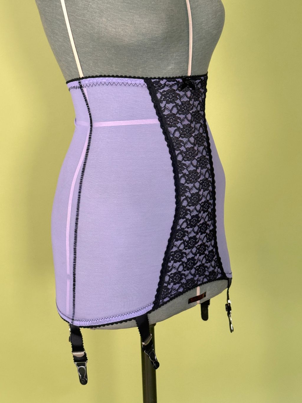 A lavender and black lace girdle with garter straps, displayed on a gray dress form mannequin against a yellow-green background.