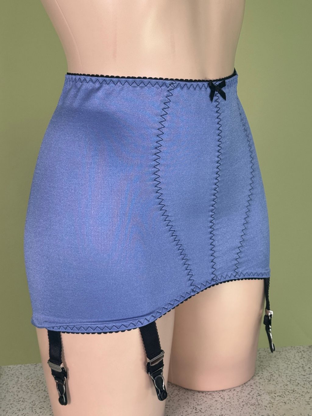 A blue girdle with black zigzag stitching, a black waistband trim, and attached black garter straps, displayed on a beige mannequin torso against a solid olive green background.
