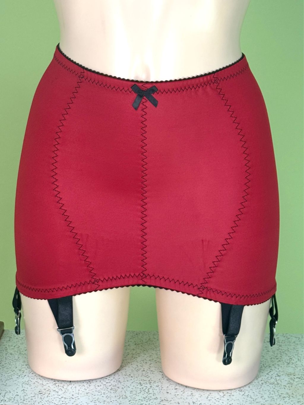 Red girdle with black zigzag stitching, a small black bow at the waistband, and attached black garter straps, displayed on a mannequin against a light green background.
