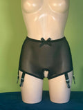 Black sheer mesh briefs with a bow at the waistband and attached garter straps, displayed on a beige mannequin torso against a green backdrop.