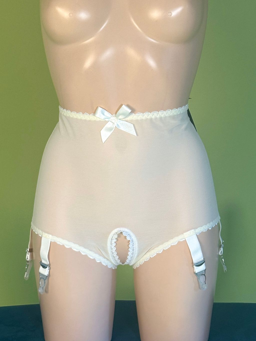 White high-waisted sheer panty with lace trim, a bow at the waist, and attached garter straps, displayed on a beige mannequin against a green background.