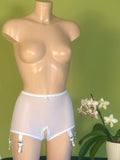 White sheer mesh garter belt and panty set with white lace trim and garter clips, displayed on a beige mannequin torso against a green wall, with a potted white orchid on the right.