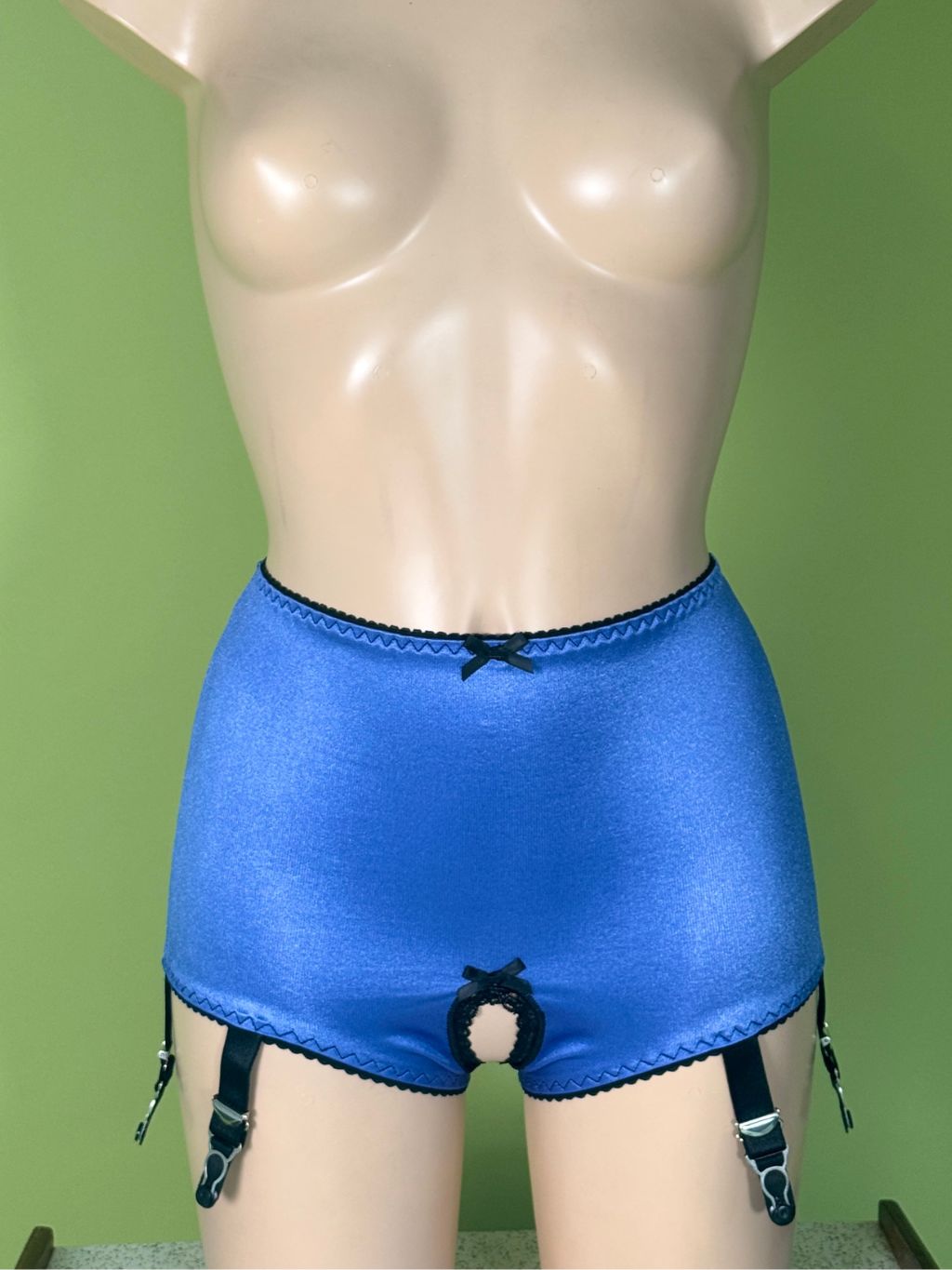 Blue satin high-waisted briefs with black trim, garter straps, and a front opening, displayed on a beige mannequin torso against a green wall.