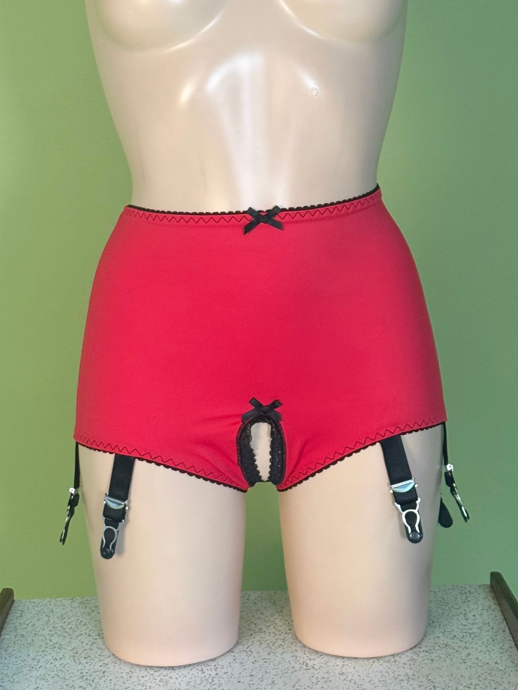 Bright red high-waisted garter belt briefs displayed on a beige mannequin torso, featuring black lace trim, a small black bow at the waistband, and attached black suspender straps with silver metal clips against a light green background.