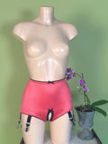 Pink garter belt and panty set with black trim and clips, displayed on a beige mannequin torso against a light green wall, with a potted orchid on the right.