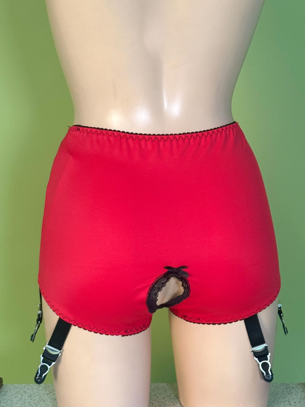 Red garter shorts with a front cutout and black suspender straps, displayed on a mannequin against a green background.