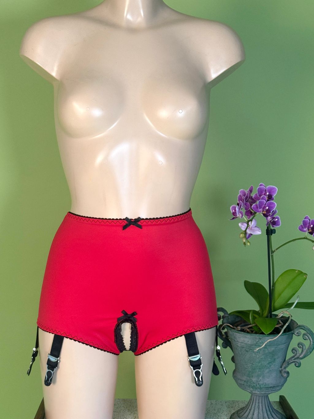 Red garter shorts with black trim and garter straps displayed on a beige mannequin torso against a green wall, with a potted purple orchid on the right.
