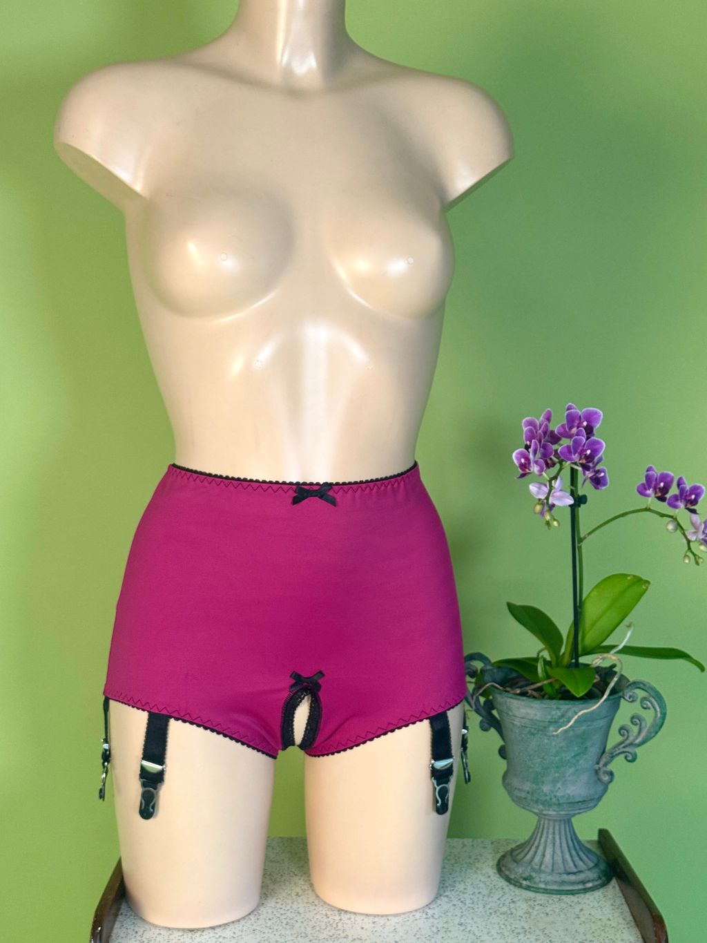 Bright pink garter shorts with black straps and bows, displayed on a beige mannequin torso against a green wall, with a potted purple orchid on the right.