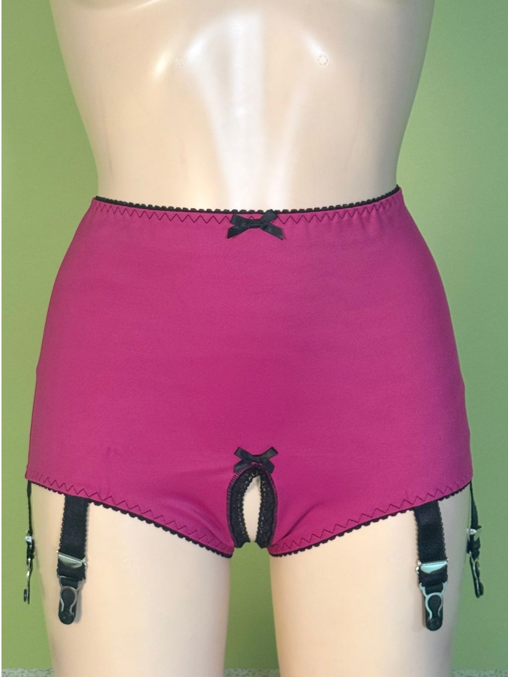 Bright pink high-waisted garter shorts with black trim, a small bow at the waist, and attached black suspender clips, displayed on a beige mannequin against a light green background.