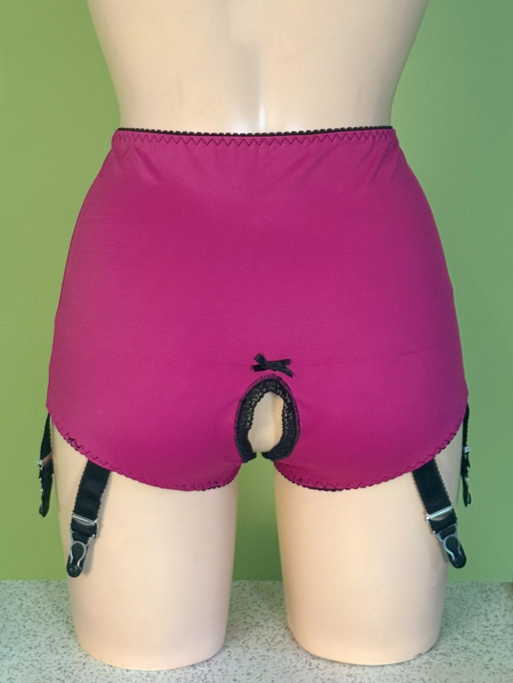 Bright pink high-waisted briefs with black lace trim and attached black garter straps, displayed on a beige mannequin against a light green background.