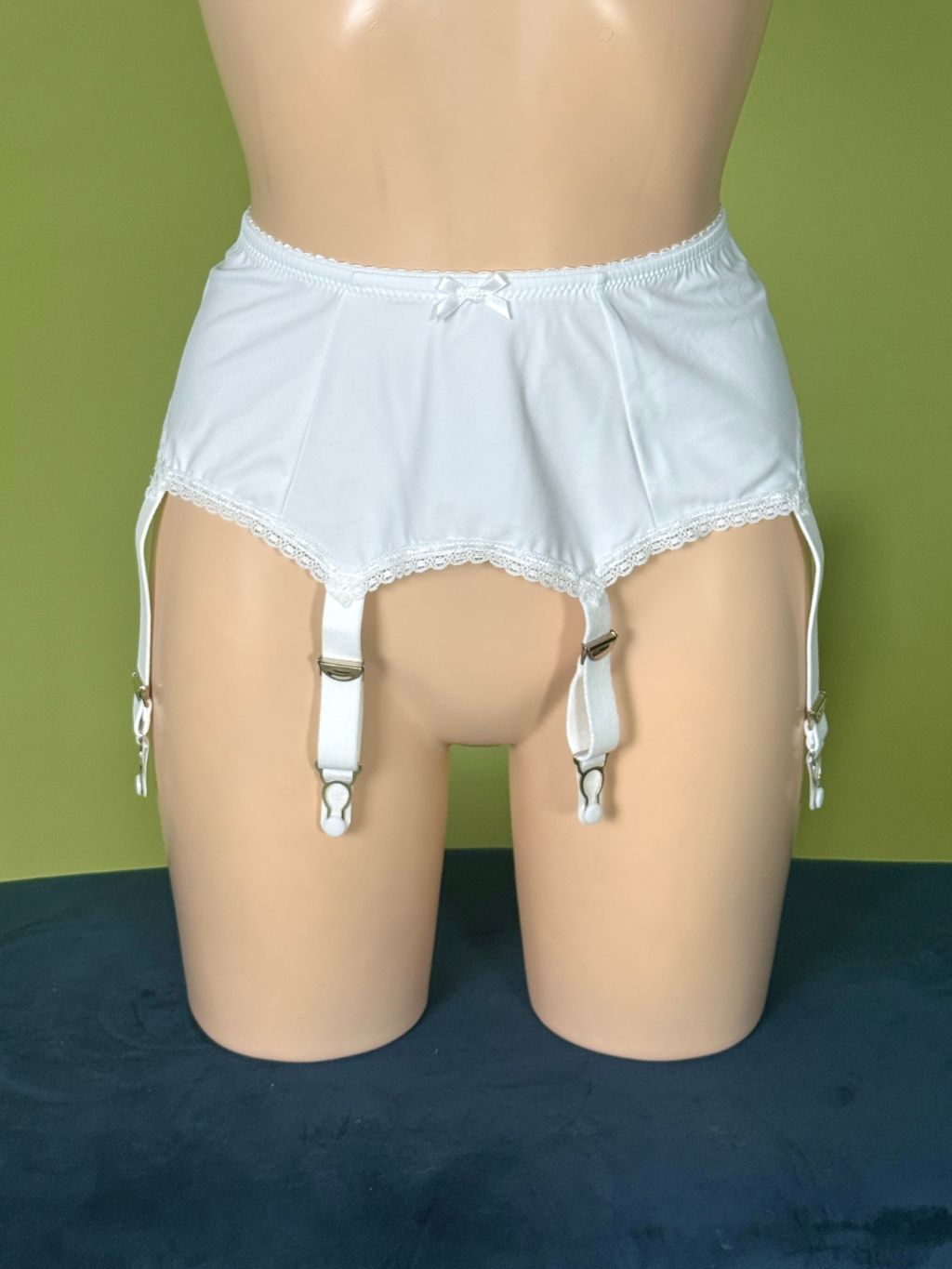 White lace-trim garter belt with attached suspender straps, displayed on a beige mannequin torso against a green wall and dark blue floor.
