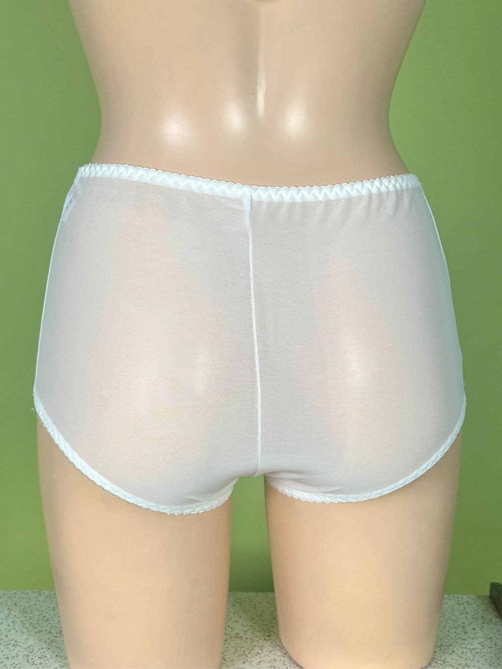 Cream sheer mesh briefs with a white braided waistband, displayed on a beige mannequin torso against a green background.