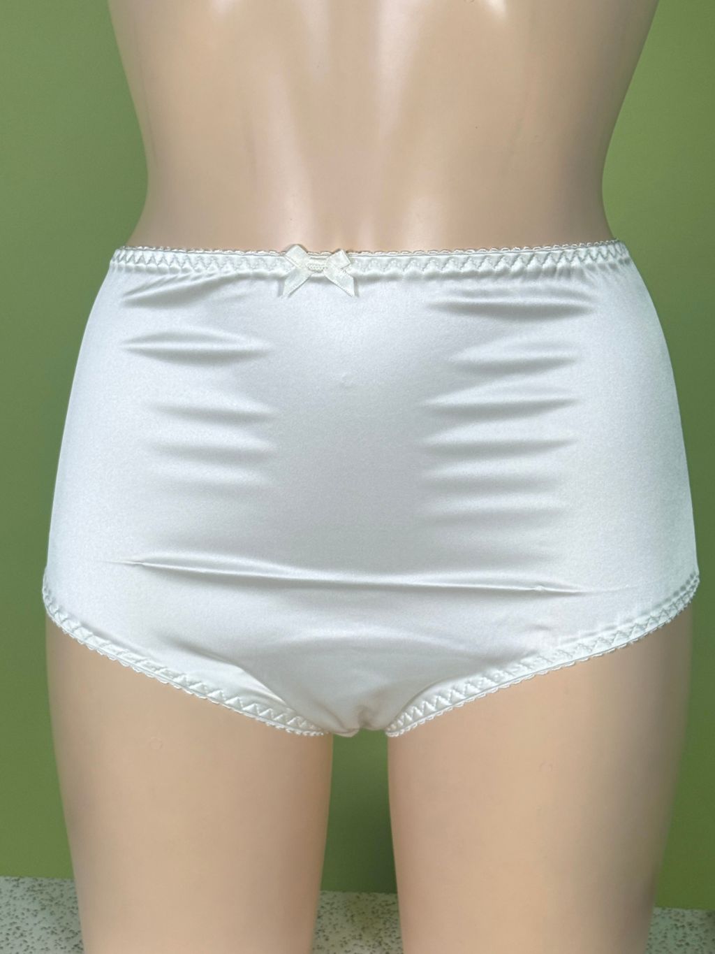 Cream satin high-waisted briefs with lace trim and a small bow at the center back, displayed on a mannequin against a green background.