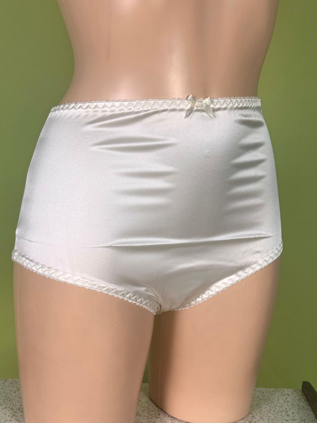 Cream satin high-waisted briefs with lace trim and a small bow at the center waistband, displayed on a mannequin torso against a green background.