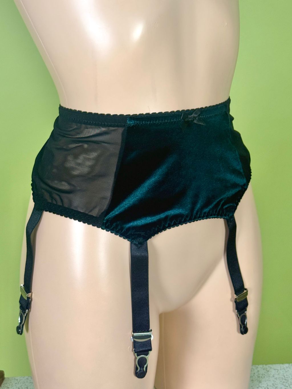 Dark green garter belt with a sheer black mesh side panel, displayed on a beige mannequin torso against a light green background.
