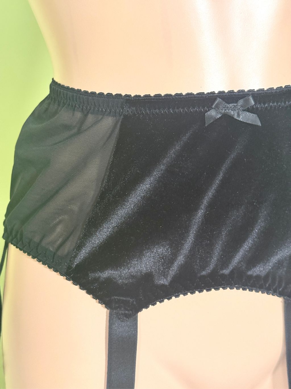 Black lingerie garter belt with sheer mesh side panels, a small bow at the front, and garter straps, displayed on a mannequin torso against a pale green background.