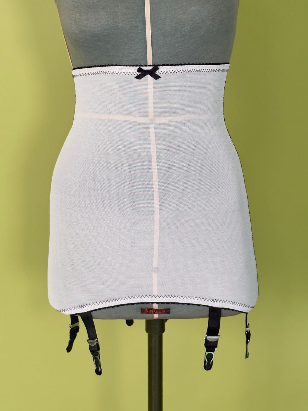 High-waisted cream girdle with black garter straps and a black bow detail, displayed on a gray dress form mannequin against a yellow-green background.
