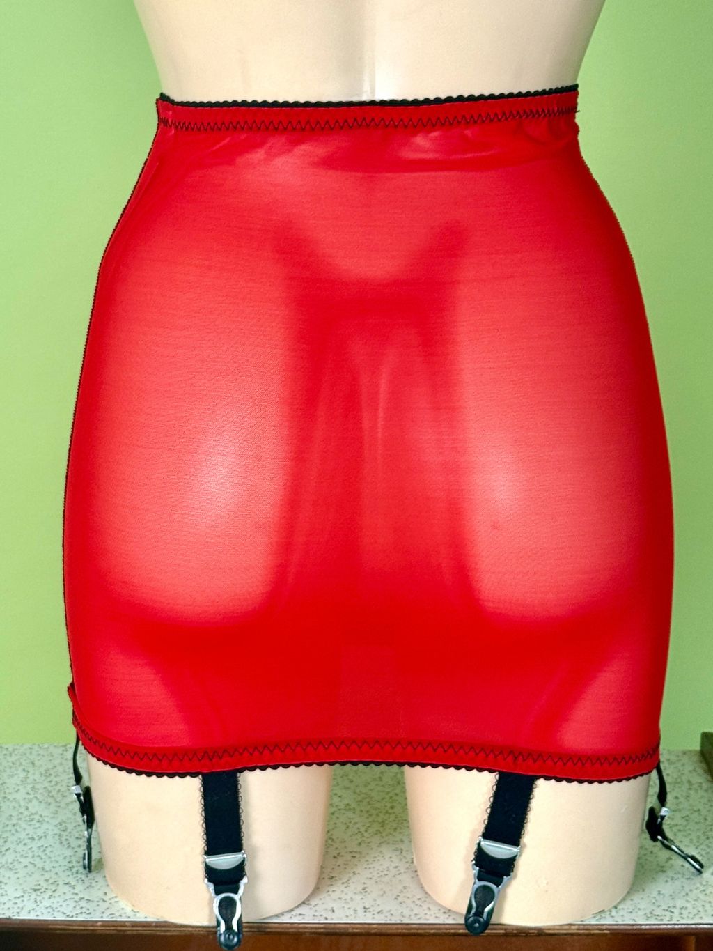 Red sheer mesh mini skirt with black trim and attached black garter straps, displayed on a beige mannequin against a light green background.