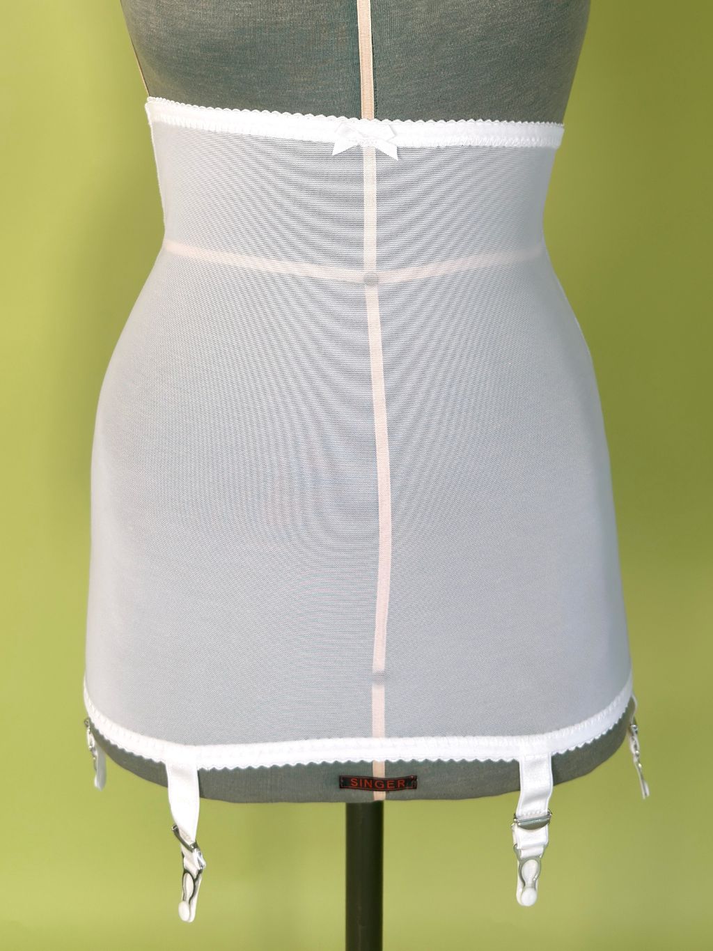 White girdle with garter straps, displayed on a gray dress form against a green background.
