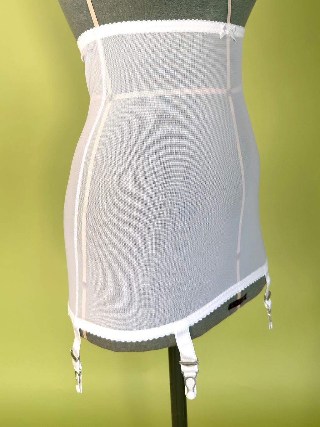 White girdle with garter straps, displayed on a dark gray mannequin torso against a solid olive green background.