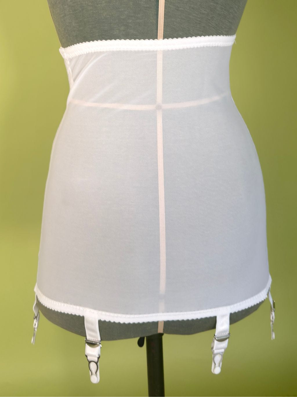 White girdle with 6 white garter clips, displayed on a gray mannequin torso against a solid olive green background.