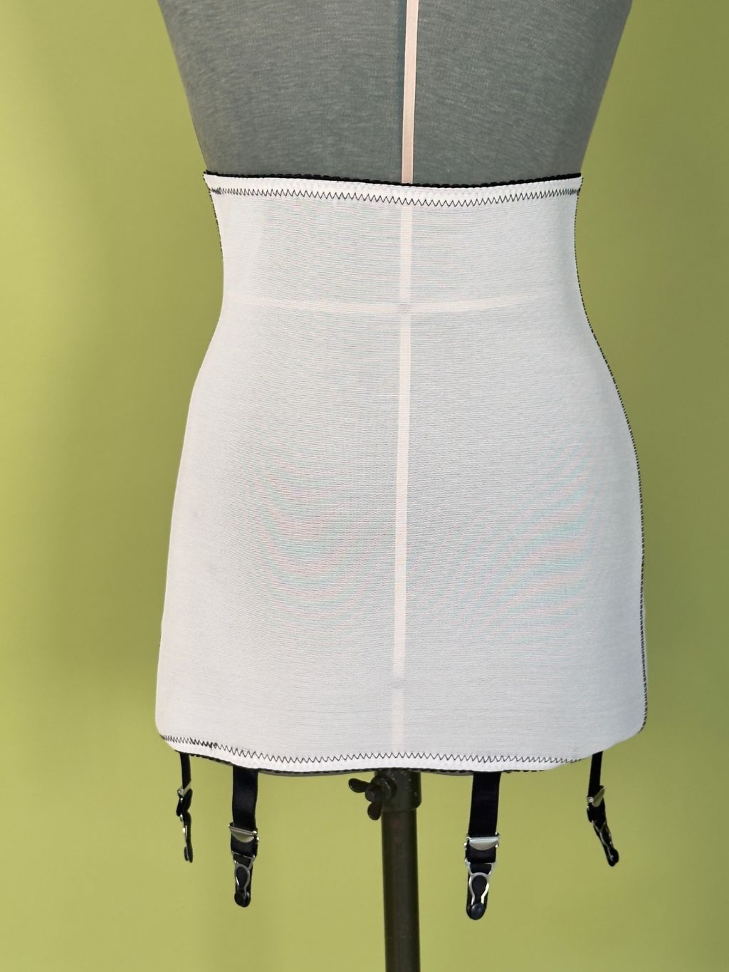 High-waisted cream girdle with black garter straps, displayed on a gray dress form against a light green background.
