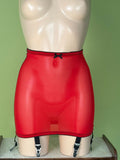 Red sheer panty with black trim, garter straps, and a small black bow at the front, displayed on a beige mannequin against a light green background.