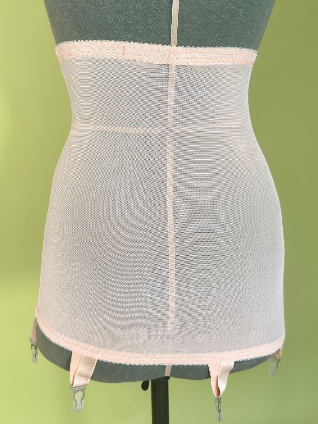 peach girdle with garter straps, displayed on a green mannequin torso against a light green background.