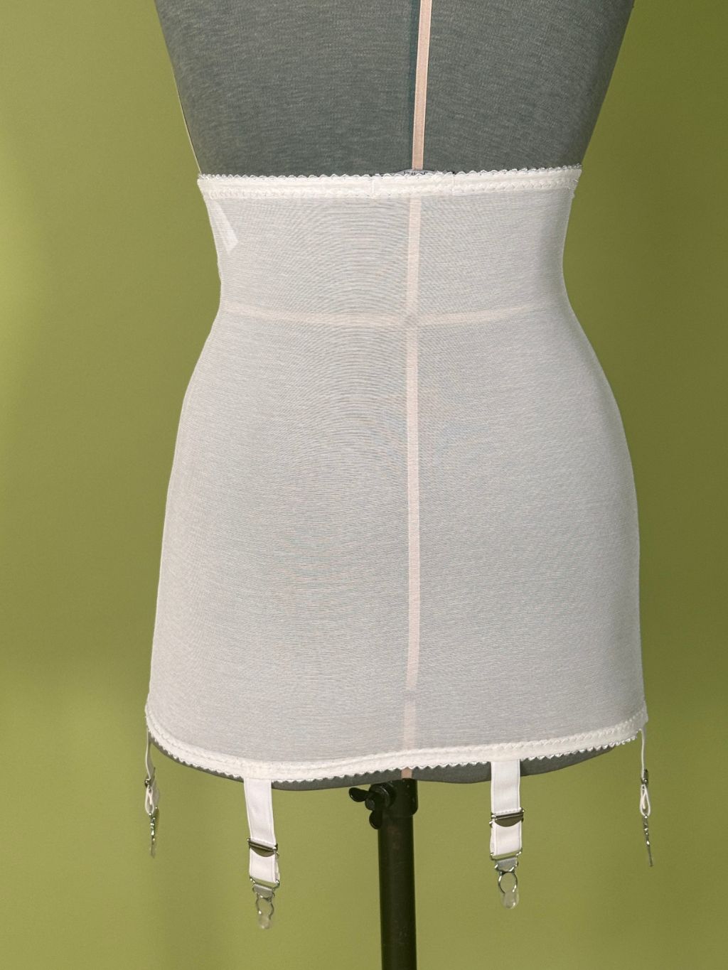 cream high waist girdle with garter straps and metal hooks, displayed on a gray dress form against a solid olive green background.