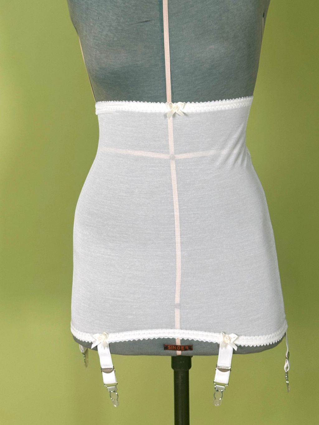 cream high waist girdle with lace trim and hanging clips, displayed on a gray dress form against a green background.