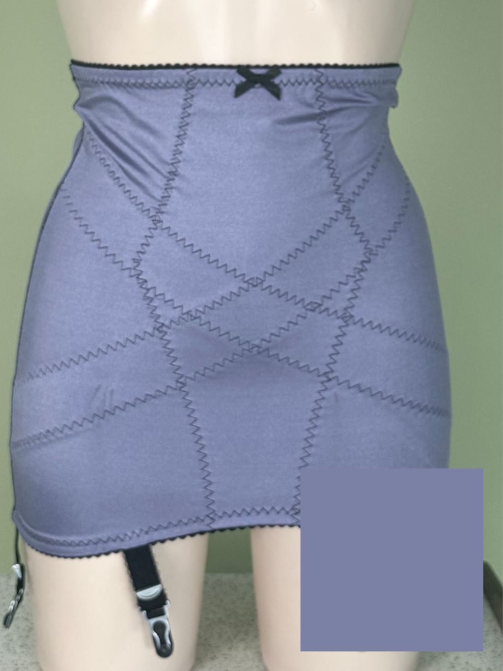 A lavender-blue high-waisted girdle with diagonal zigzag stitching, a black lace-up bow at the waistband, and garter straps on the hips, shown on a mannequin against a light green wall.