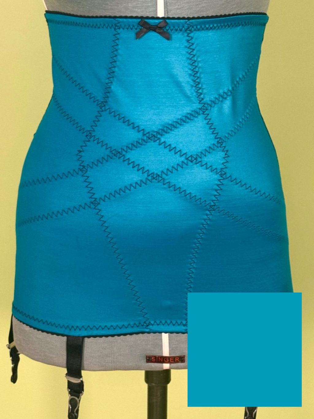 A bright turquoise blue girdle displayed on a gray dress form mannequin against a pale yellow background. The garment features a diamond argyle-style stitched pattern, a small black bow at the waistband, and garter straps hanging from the bottom edge.
