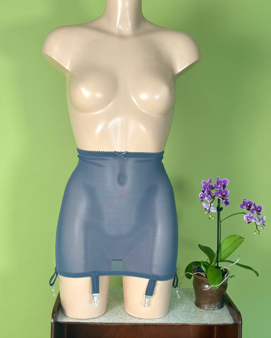 A blue-gray sheer girdle displayed on a beige mannequin torso, with a small potted purple orchid on a wooden surface against a light green background.