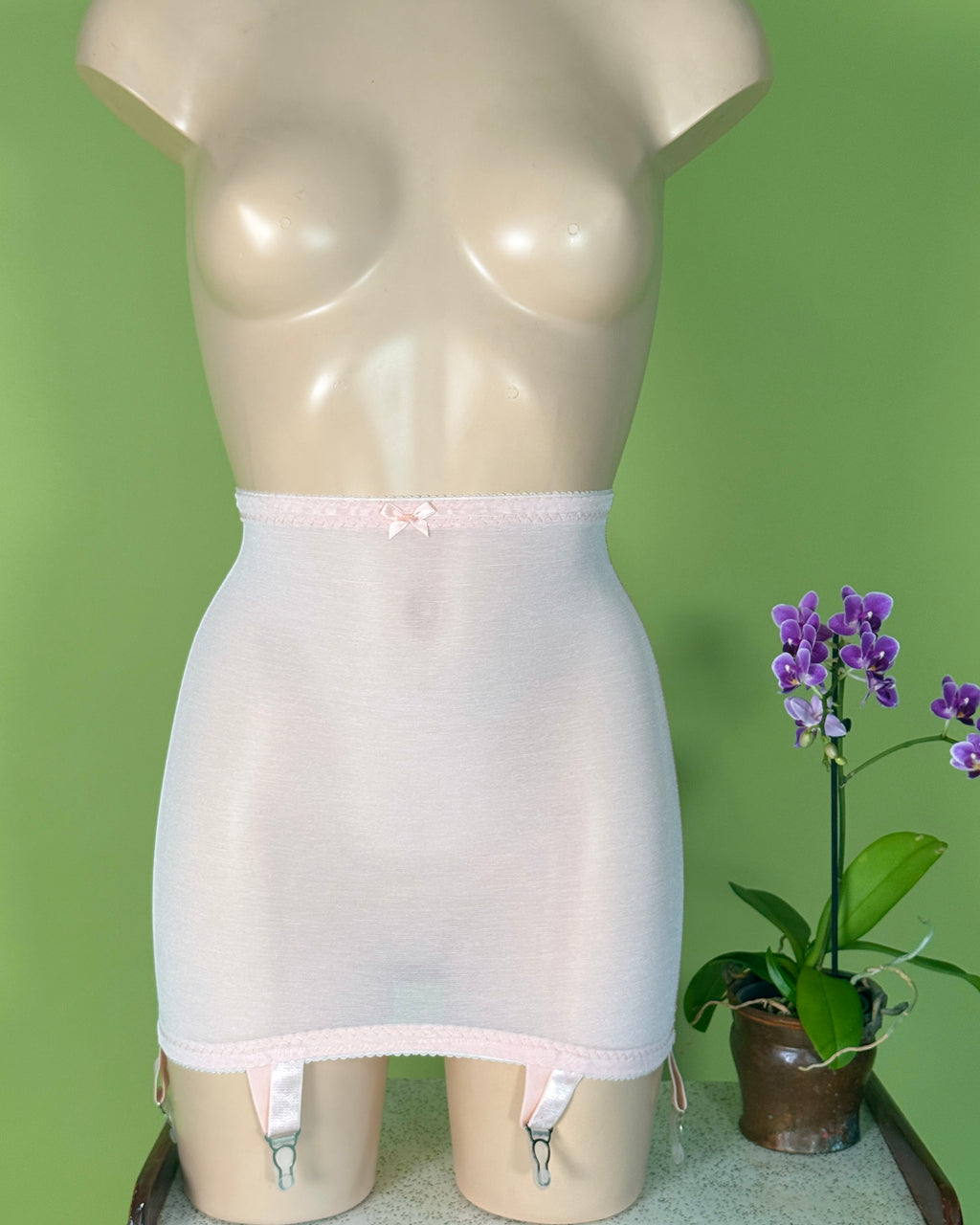 A mdisplay a light pink sheer girdle with a small bow at the waistband and attached garter straps. 